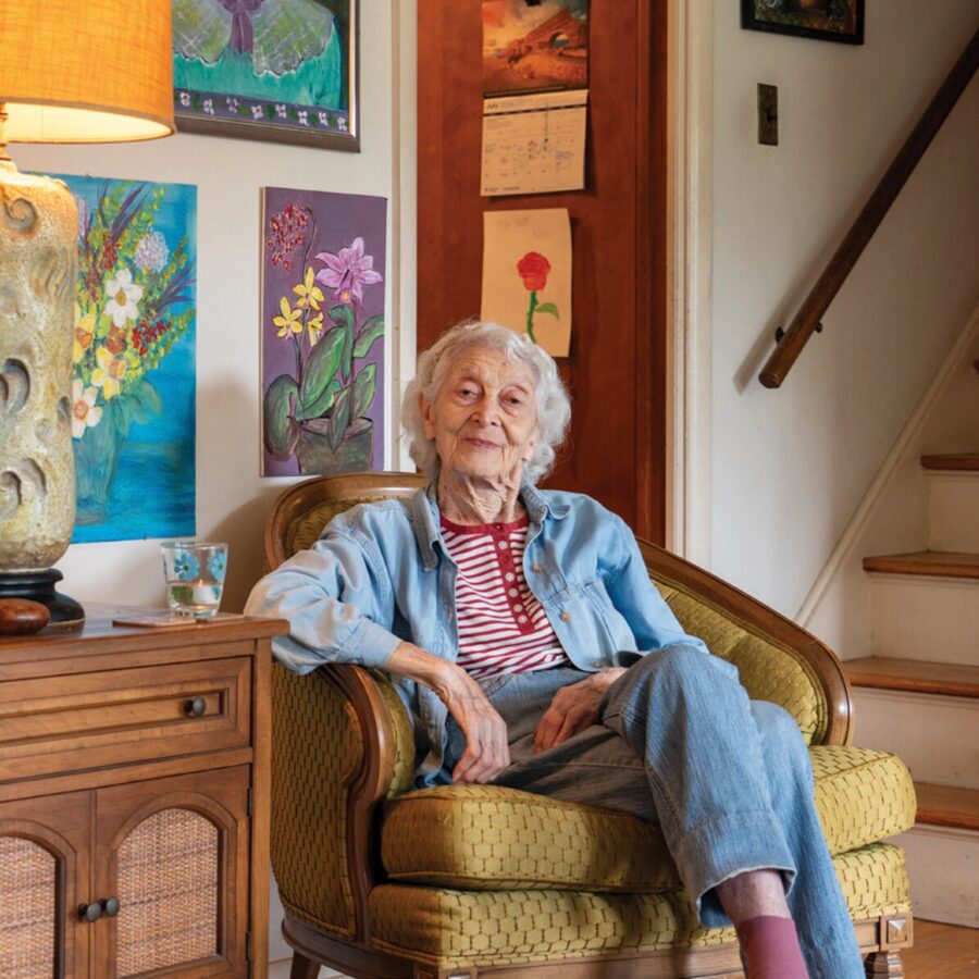 Naomi A. Reich seated in an armchair at home, surrounded by her paintings